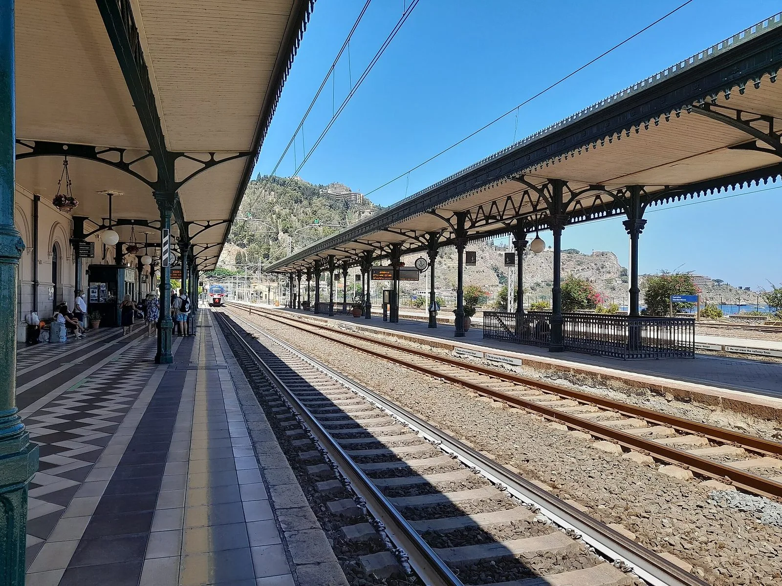 Banchina della stazione di Taormina-Giardini con le pensiline in ghisa verde, i binari e il pavimento a scacchi, qualche viaggiatore in lontananza e la collina di Taormina sullo sfondo sotto un cielo blu