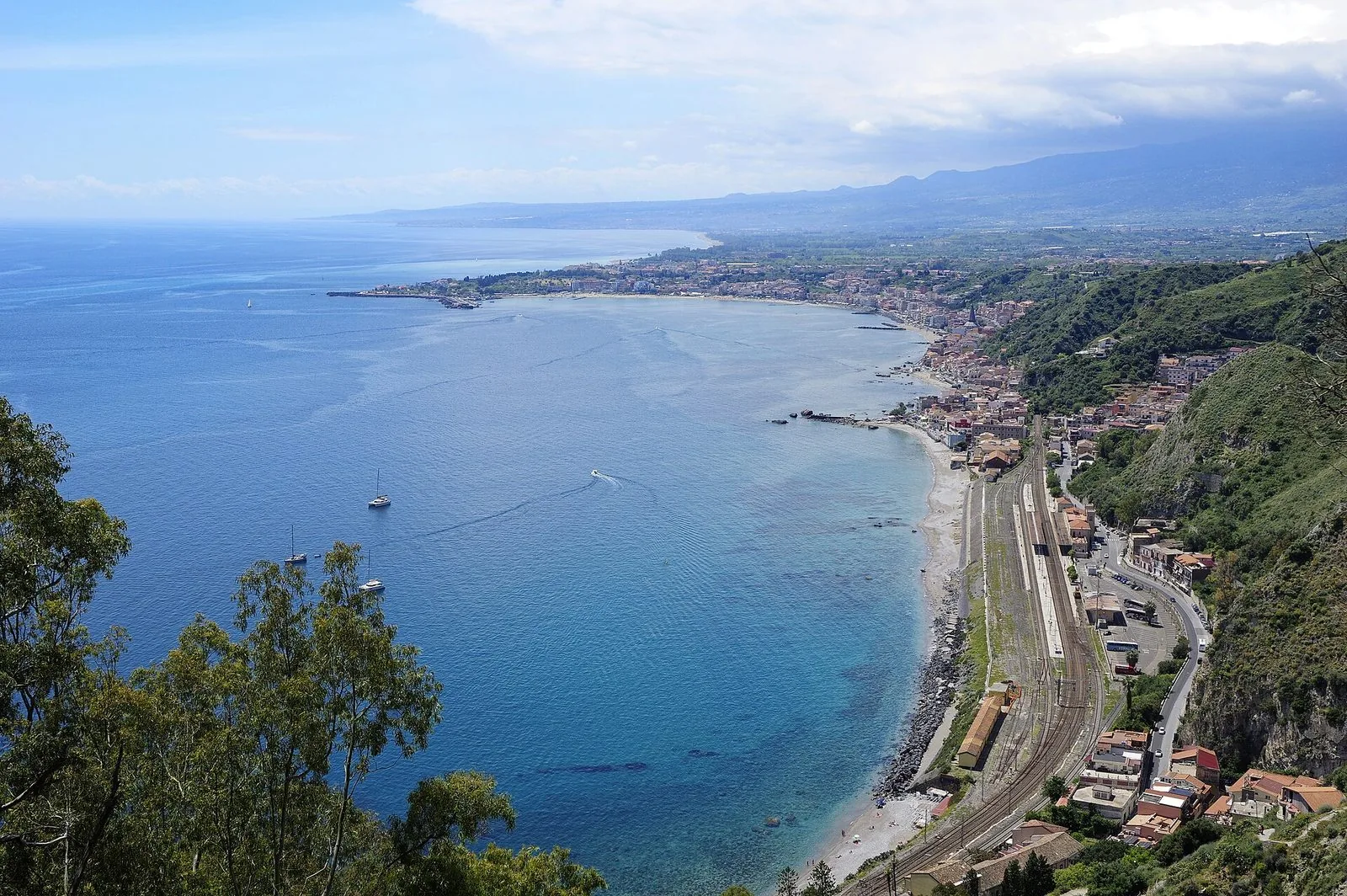 Veduta dall'alto della baia di Giardini Naxos: il mare blu, la spiaggia stretta lungo la ferrovia e la stazione, il paese disteso fino al promontorio di Capo Schisò con il molo del porto, e le colline verso l'Etna sullo sfondo