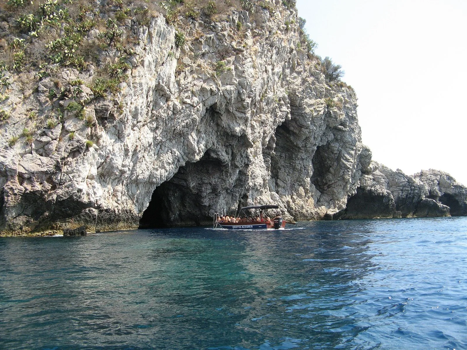 Una barca turistica con passeggeri all'ingresso di una grotta marina scavata in una scogliera calcarea chiara, con fichi d'India sulla parete e mare blu e trasparente in primo piano