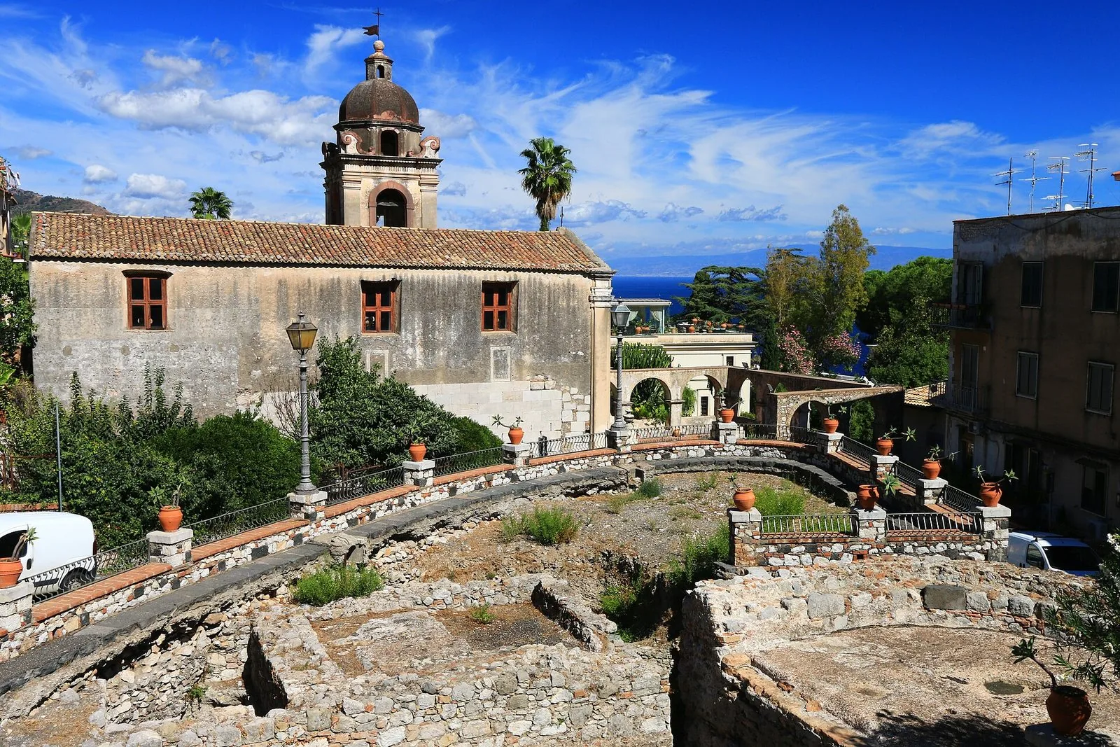 La chiesa di San Pancrazio a Taormina con il campanile a cupola e il tetto in coppi, vista dall'alto sopra i resti in pietra di antiche strutture, con palme, vasi di terracotta e uno scorcio di mare blu sotto un cielo con nuvole leggere