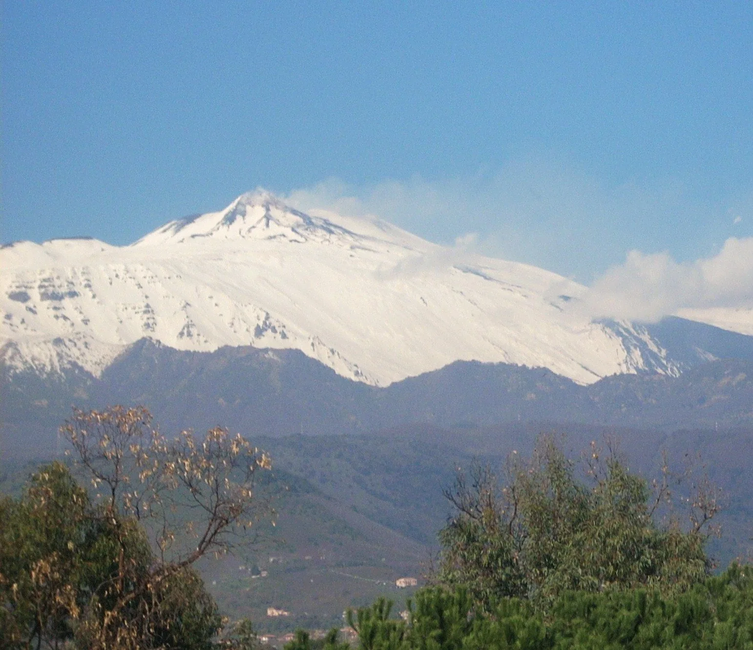 La cima dell'Etna completamente coperta di neve sotto un cielo azzurro, con nuvole basse sul fianco destro e colline verdi e alberi in primo piano