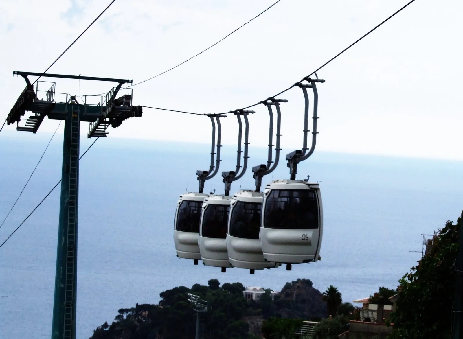 Quattro cabine bianche della funivia di Taormina sospese ai cavi accanto a un pilone verde, con il mare Ionio e un promontorio alberato sullo sfondo