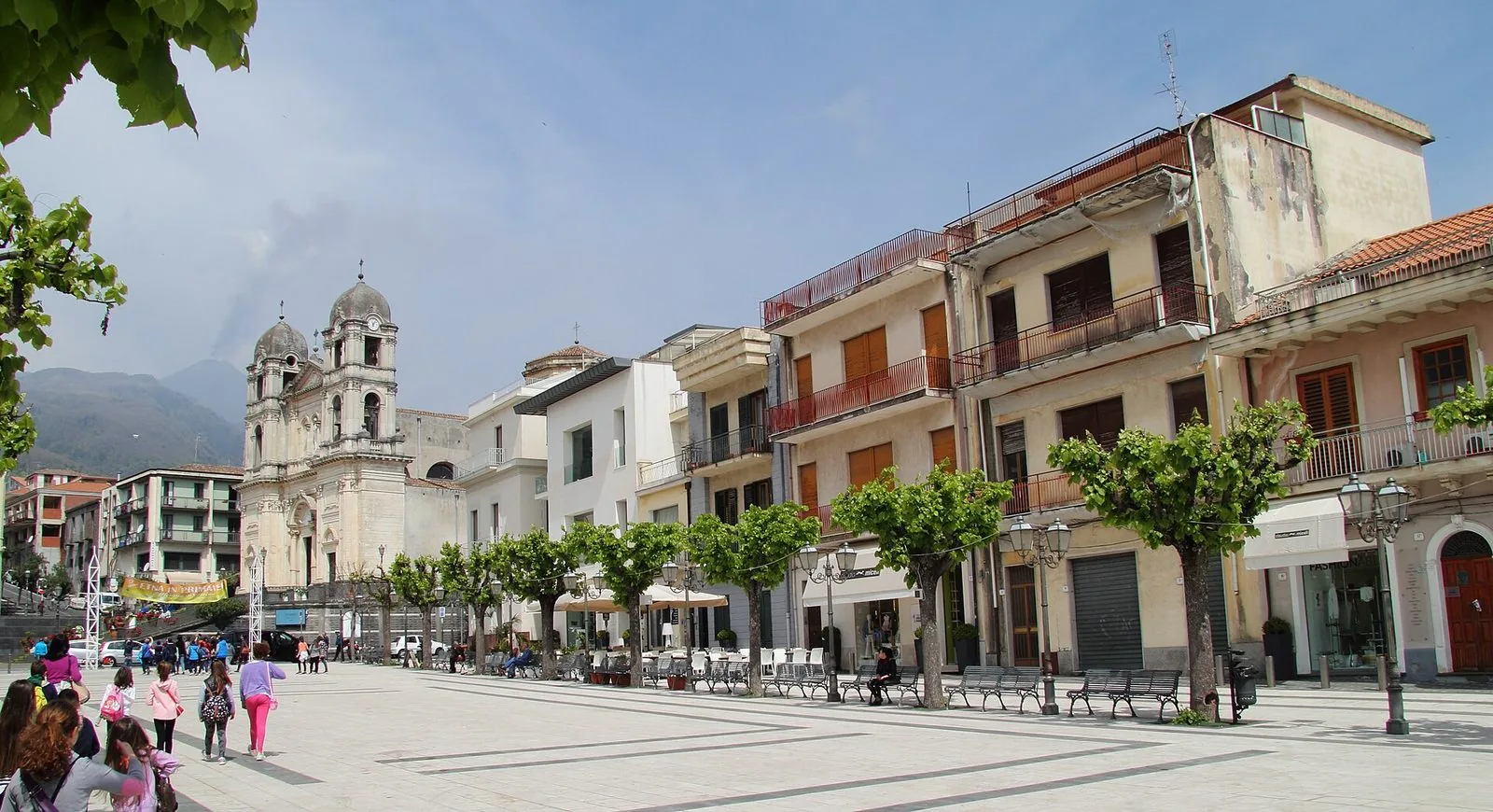 La piazza principale di Zafferana Etnea con la facciata in pietra chiara della chiesa madre a due campanili, una fila di platani potati, panchine e palazzi a più piani, con le pendici dell'Etna e un pennacchio di fumo sullo sfondo