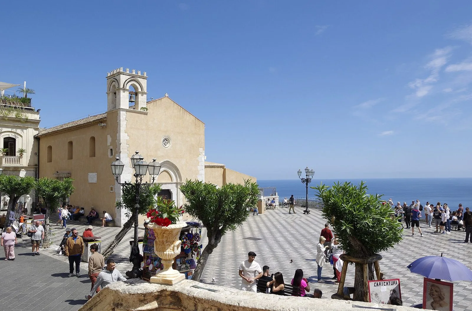 Piazza IX Aprile a Taormina in una giornata di sole: la facciata in pietra chiara della chiesa di Sant'Agostino con il campanile merlato, la pavimentazione a scacchi bianchi e grigi, alberelli, lampioni in ferro e il belvedere aperto sul mare blu