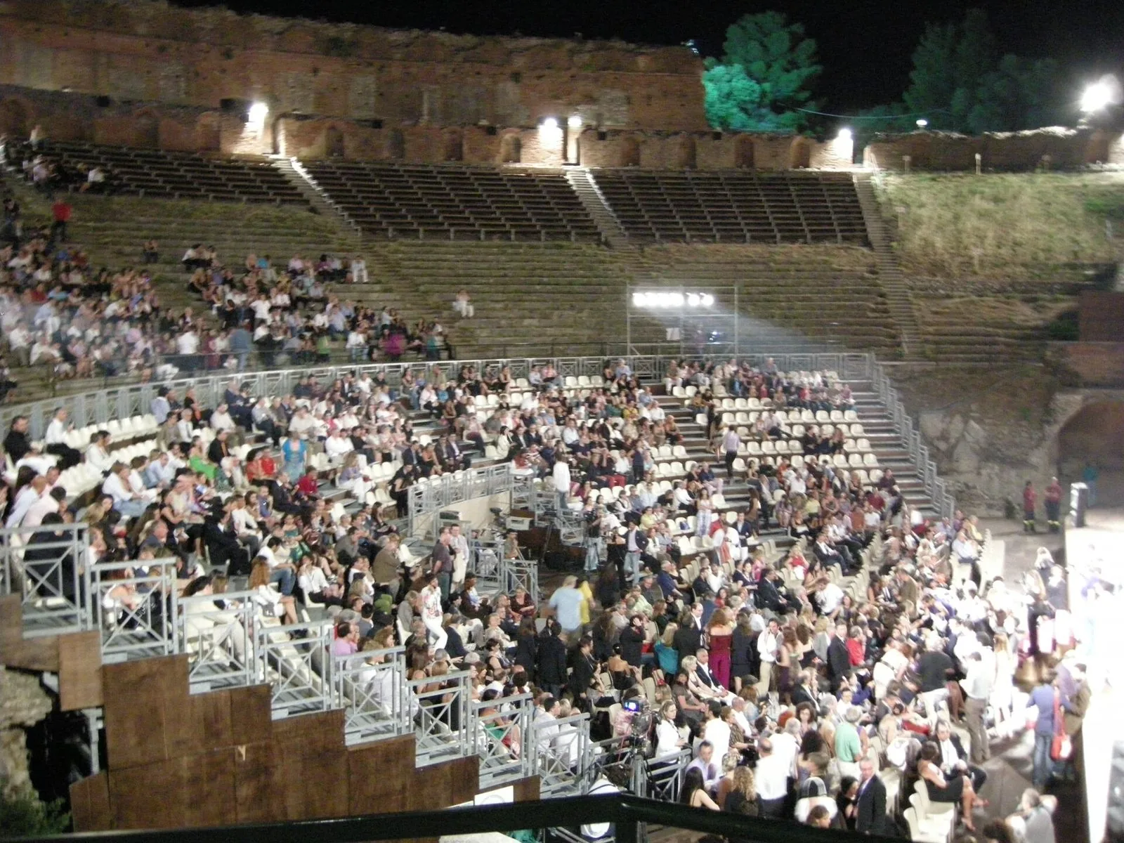 La cavea del Teatro Antico di Taormina di notte durante il Taormina Film Fest: gradinate e tribune piene di spettatori illuminate dai riflettori, con i muri in mattoni dell'antica scena sullo sfondo